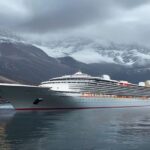 A photorealistic image of a large cruise ship docked at the port of Ushuaia, Argentina, with snow-capped mountains in the background. The ship's name is not visible. The scene is overcast, conveying a