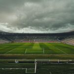 A realistic photo of the Eduardo Gallardón stadium in Santa Fe, Argentina, during a football match. The pitch is small, with worn grass, and the stands are full of local fans. The sky is overcast, an