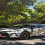 A photorealistic image of a 2022 Toyota Corolla Hybrid in silver parked in a suburban driveway, with a 'For Sale' sign on the windshield, sunny day, green trees in background.