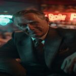 A photorealistic movie still of Glen Powell as a charismatic con artist in a dimly lit New York bar, wearing a tailored suit, leaning casually against a wooden counter, smirking at the camera, with bl