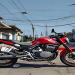 A photorealistic image of a new red Motomel Blitz One Base 110cc motorcycle parked on a street in a typical Argentine neighborhood, with a price tag on the handlebar showing ARS 1,200,000. The backgro
