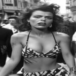 A black and white photograph of Valie Export in the 1960s, wearing a provocative outfit, performing a body art piece in a public street, with a confrontational expression, surrounded by a crowd of onl