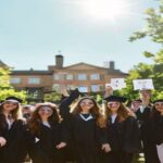 A group of diverse young graduates in caps and gowns celebrating outdoors in front of a university building in Galicia, holding diplomas, with a sunny sky and green campus in the background.