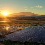 A photorealistic wide shot of a solar panel field in Sonora, Mexico, with the sun setting behind the panels, casting long shadows. In the background, a traditional Mexican village with adobe houses an