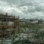 A photorealistic image of an unfinished hospital construction site in a rural area of Honduras, with rusted scaffolding and overgrown weeds, symbolizing abandoned projects and cost overruns. The backg