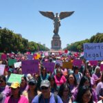A photorealistic image of a large women's march in Mexico City, with colorful banners reading 'Ni una más' and 'Aborto legal', thousands of women wearing purple and green, in front of the Angel of In