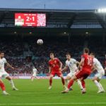 Photorealistic image of a football match between Hungary and Finland in a large stadium, with Hungarian players in red and white attacking and Finnish players in white defending. The scoreboard shows
