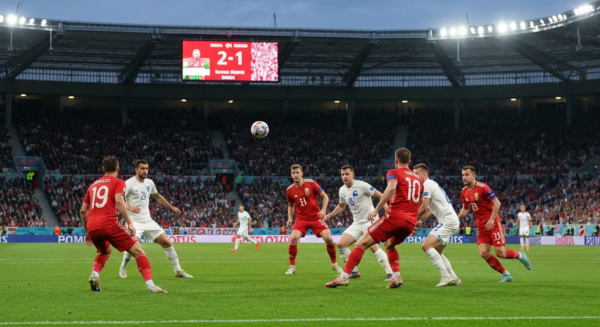 Photorealistic image of a football match between Hungary and Finland in a large stadium, with Hungarian players in red and white attacking and Finnish players in white defending. The scoreboard shows