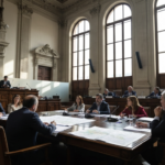A photorealistic image of a legislative assembly hall in San Juan, Argentina, with lawmakers debating around a large wooden table, documents and maps spread out, natural light streaming through tall w