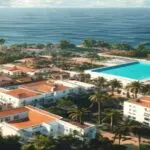 A photorealistic aerial view of a modern residential complex in the Canary Islands, with white buildings, terracotta roofs, and a swimming pool, surrounded by palm trees and the Atlantic Ocean in the