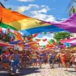 A vibrant Pride parade in a small Spanish town, with colorful rainbow flags, diverse people of all ages smiling and marching, banners with messages of love and respect, sunny day, cobblestone streets,